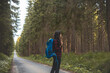 © Fauren - Back view of a woman with a blue backpack walking barefoot down a peaceful forest road, surrounded by lush greenery and tall trees, symbolizing adventure and tranquility in nature