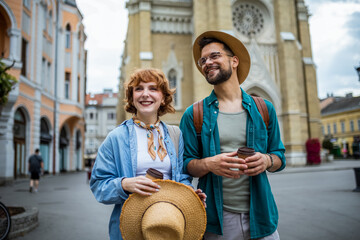 Wall Mural - Young couple walking through the new city on their vacation. They are drinking coffee and looking at monuments and buildings.