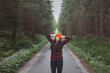 © Fauren - Smiling woman in a red checkered shirt and bright orange beanie stands in the middle of a forest road, stretching her arms behind her head, enjoying the tranquility of the surroundings