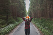 © Fauren - Smiling woman in a red checkered shirt and bright orange beanie stands in the middle of a forest road, stretching her arms behind her head, enjoying the tranquility of the surroundings