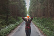 © Fauren - Smiling woman in a red checkered shirt and bright orange beanie stands in the middle of a forest road, stretching her arms behind her head, enjoying the tranquility of the surroundings