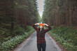 © Fauren - Smiling woman in a red checkered shirt and bright orange beanie stands in the middle of a forest road, stretching her arms behind her head, enjoying the tranquility of the surroundings