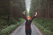 © Fauren - Smiling woman in a red checkered shirt and bright orange beanie stands in the middle of a forest road, stretching her arms behind her head, enjoying the tranquility of the surroundings