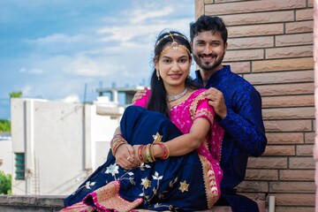 Young beautiful Indian couple in traditional Rajasthani, Gujarati Indian attire sitting on terrace for a pre wedding celebration, engagement, photoshoot, ring ceremony, festival, teej, love, marriage.