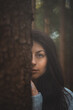 © Fauren - Enigmatic woman with captivating blue eyes peeks from behind a tree in a mystical forest. The image captures the essence of mystery, nature, and serene beauty