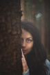 © Fauren - Enigmatic woman with captivating blue eyes peeks from behind a tree in a mystical forest. The image captures the essence of mystery, nature, and serene beauty