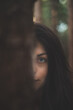 © Fauren - Enigmatic woman with captivating blue eyes peeks from behind a tree in a mystical forest. The image captures the essence of mystery, nature, and serene beauty