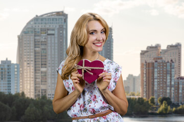 Portrait of happy smiling mature blonde woman holding a red heart shape gift box in both hands. Residential area buildings in the background.