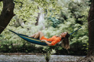 Woman in an orange dress relaxing in a hammock by a forest stream, smiling and enjoying the peaceful natural surroundings.