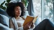 © Emilio - Young black woman enjoying me-time at home. African american adult girl reading book on couch in her living room. Tranquil and relaxing solo hobby
