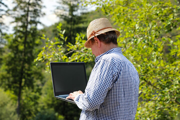 Programmer in straw hat and checkered shirt standing in forest high in mountains working on laptop. Digital Nomads series.