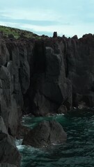 Wall Mural - Top view of black rocks with blue waves on cloudy day. Clip. Black stone cliffs on ocean shore on cloudy day. Northern coasts on cloudy summer days