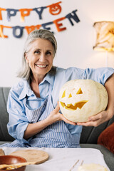 Wall Mural - Senior, smiling woman holding carved pumpkin, looking at camera celebrating halloween at home