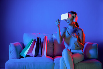 Wall Mural - Smart female sit on sofa beside bags wear VR headset connecting metaverse, future cyberspace community technology. Elegant woman enjoy shopping products from online store in meta world. Hallucination.