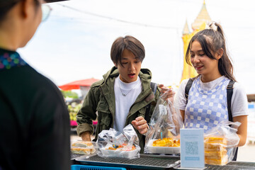 Wall Mural - Young Asian man and woman buying and eating street food at railway station with using mobile app on smartphone scan QR code making online payment during travel local town on summer holiday vacation