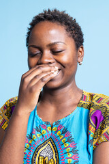 Wall Mural - Joyful african american model standing over blue background, posing with confidence while dressed in traditional ethnic costume in studio. Feminine gorgeous woman laughing on camera.