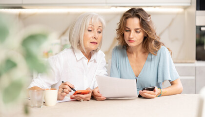 Mother and daughter sitting at table and calculating finances. Daughter using her smartphone for online payment