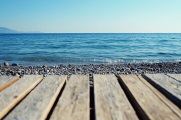 A tranquil seascape featuring a calm blue ocean meeting a pebbled shore, viewed from a wooden pier under a clear sky.