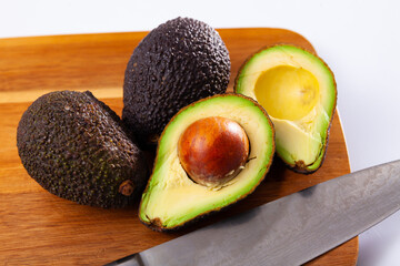 Whole and sliced avocados on wooden board with kitchen knife, over white background.