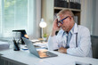 © Wasana - Senior doctor experiencing burnout sitting at his desk looking worried at laptop screen