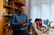 © DusanJelicic - Business man in a blue shirt smiling while looking at his smart phone in an office setting. In the background, two colleagues are working at a desk with laptops.