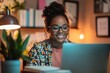 © FarisZul - A young Black woman is smiling and working on her laptop in the evening at home, sitting near a desk with a bookcase behind her.