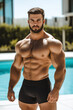 © fotogurmespb - A muscular Caucasian man in black swim trunks poses confidently by a pool, representing fitness and summer vacation