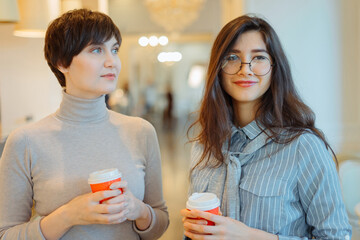 Two young smiling women standing in cafe holding hot coffee in paper cup