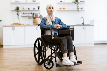 Muslim woman wearing hijab sitting in wheelchair at home in kitchen with modern decor. Woman in casual clothes looks calm and confident. Inclusive lifestyle independence and accessibility concept.