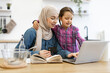 © sofiko14 - Happy family cooking together, embracing technology for home activities. Muslim mother and daughter sitting at kitchen table and buying ingredients for meal using laptop, bank card and recipe book.