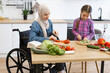 © sofiko14 - Muslim mother in wheelchair and daughter cutting fresh vegetables for salad in modern kitchen. Enjoying family time, cooking together. Inclusive, love, and nurturing relationship