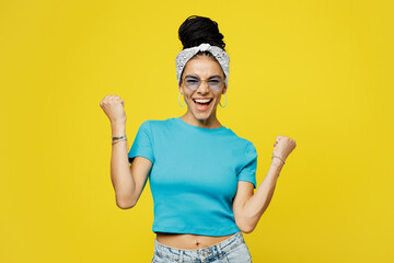 Wall Mural - Young excited happy cool Latin woman she wearing blue top t-shirt headband glasses casual clothes do winner gesture clench fist isolated on plain yellow background studio portrait. Lifestyle concept.