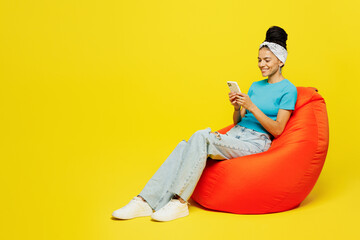 Wall Mural - Full body young happy Latin woman she wears blue top t-shirt headband casual clothes sit in bag chair hold in hand use mobile cell phone isolated on plain yellow background studio. Lifestyle concept.