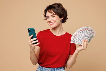 Wall Mural - Young surprised shocked woman she wear red t-shirt casual clothes use mobile cell phone hold in hand fan of cash money in dollar banknotes isolated on plain pastel beige background. Lifestyle concept.