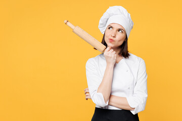 Wall Mural - Young minded pensive chef cook baker woman wear white shirt black apron uniform toque chefs hat hold in hand rolling pin look aside on area isolated on plain yellow background. Cooking food concept.