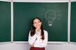 © deagreez - Photo of charming teen pupil girl thinking electric bulb idea wear trendy uniform classroom blackboard background