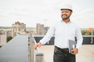 Asian Indian male contractor engineer with hard hat standing in front construction site, inspecting the progress