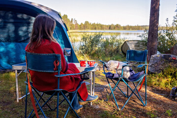 woman, sitting on a camping chair in front of a tent, enjoying cup of coffee and amazing view in nor