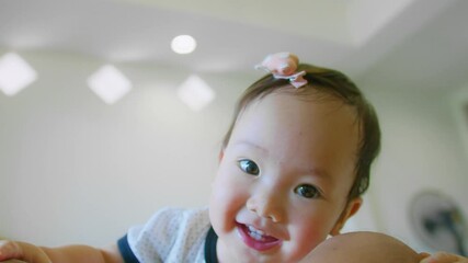 Poster - Adorable Cute Asian baby toddler with a bow on head smiling and looking up at the camera in a bright room with soft lighting.