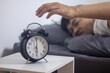 © vachiravit - The man lies in bed, reaching out to his alarm clock.