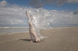 © Milou Dirks - art portrait of woman in off white dress standing in the wind on the beach holding thin plastic sheet in sun light