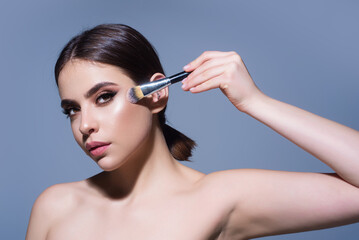 Wall Mural - Beautiful young woman applying makeup on studio background. Pretty girl holding makeup brushes and make up on face with cosmetics. Facial beauty. Perfect skin, natural make up.