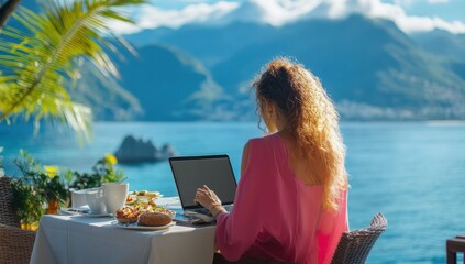 Poster - A woman in a pink dress works on a laptop at a breakfast table overlooking the mountains and sea from a restaurant terrace at a luxury hotel. The woman has long curly hair