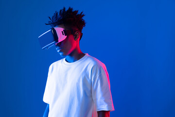 Surprised young African American looking through VR holding interesting object hologram on pink blue neon wall at metaverse world connecting digital futuristic technology virtual reality. Contrivance.