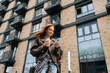 © dikushin - Low-angle view of stylish cheerful young woman standing on city street while drinking coffee and browsing smartphone with smile on face, posing on blurred background of modern building.