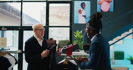 Wall Mural - Regular elderly client asking store worker for assistance with clothing items, looking for a shirt to go with his suit jacket. African american clerk managing inventory and helping shopper. Camera B.