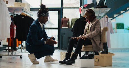 Wall Mural - Retail clerk recommending pair of boots to pregnant woman, offering assistance to find the perfect size. African american client trying on footwear on sale at clothing store, consumerism. Camera B.