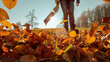© helivideo - LOW ANGLE VIEW, LENS FLARE, DOF: Colorful fallen leaves blown by a leaf blower. A scattering of flying autumn foliage with an unrecognizable woman using a portable gardening device in the background.