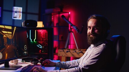 Wall Mural - Portrait of smiling man in dark living room playing engaging video games on gaming PC at computer desk, chilling after work. Happy gamer contending against foes in online multiplayer shooter