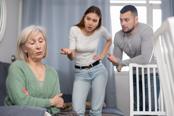 Wall Mural - Sad mature woman sitting at home while emotional young adult couple berating her, family relationship problems concept